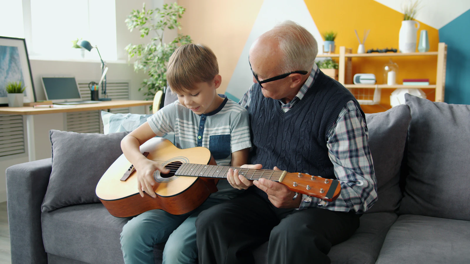 A grandfather helping his grandson learn to play guitar on the couch at home